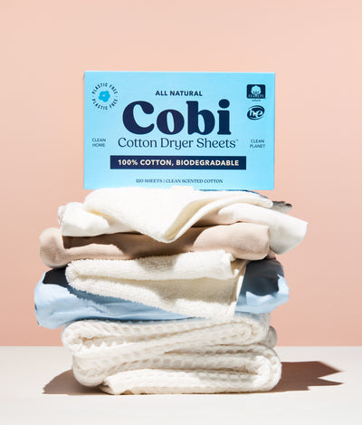cobi dryer sheet box, scented, on top of folded laundry, pink background.